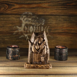 May include: A wooden sculpture of a robed figure holding a sword and a mask, with the inscription "Nemxin" at the base. Two small, dark candle holders flank the sculpture. The background is a wooden panel with a tree design.