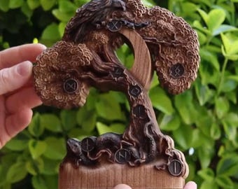 May include: A detailed wooden sculpture of a tree with intricate carvings. The sculpture features a bird at the top, runes, and a serpent at the base. The tree is brown with a natural wood grain finish, set against a green background.