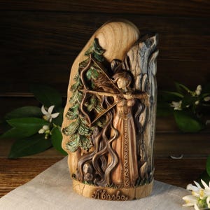 May include: A wooden carving of a woman with flowing hair, holding a bow and arrow, standing in front of a tree. The carving is painted in shades of brown, green, and orange. The text "Alnboða" is carved at the bottom of the piece.