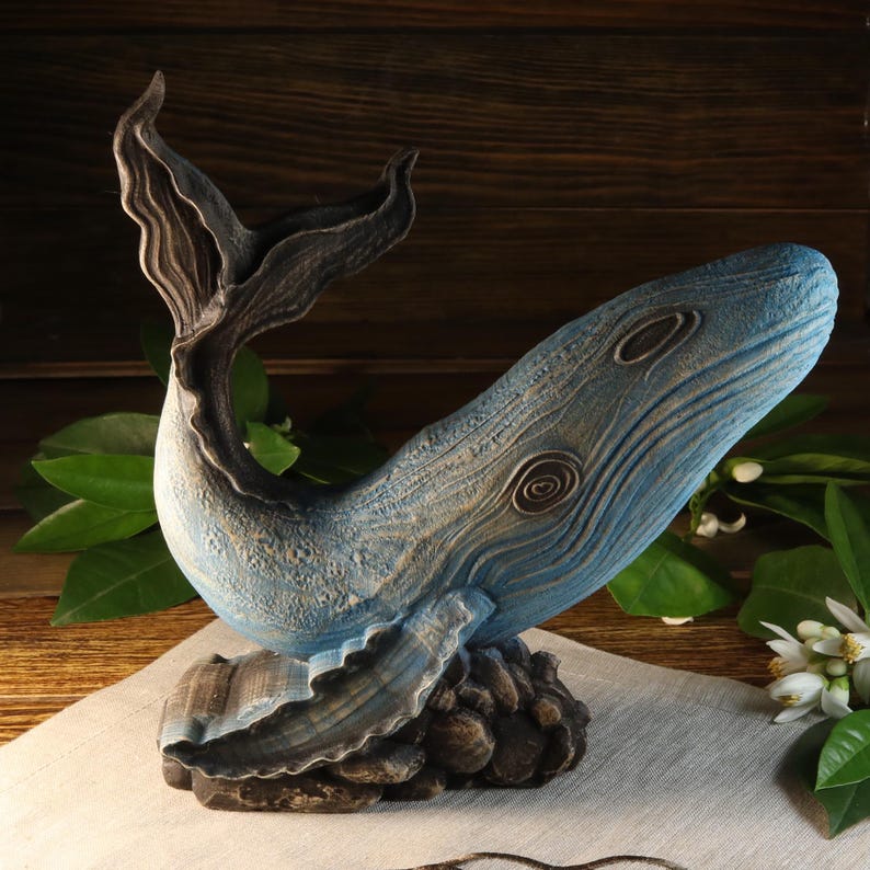 A blue whale sculpture with a textured surface and detailed fins and tail. The whale is resting on a dark, textured base. The sculpture has a light blue body with darker blue accents and is set against a wooden background.