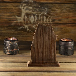May include: A wooden, abstract-shaped display stand with a dark brown finish. Two lit candles in dark, textured holders flank the stand. The background features a wooden panel with the word "NORHUR" and a tree design.