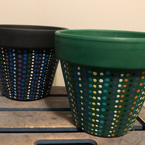 May include: Two terracotta flower pots with polka dot patterns. One pot is black with blue, purple, green, and white dots. The other pot is green with yellow, orange, white, blue, and purple dots.