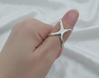 May include: A silver-colored ring with a cross-shaped design. The ring is worn on a thumb and has a modern, minimalist aesthetic. The ring's smooth, polished surface reflects light, creating a bright appearance.