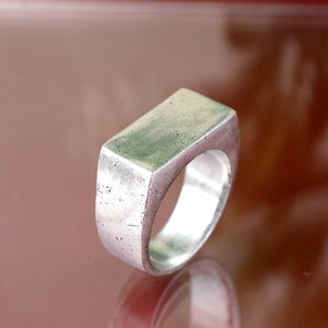 Può includere: Anello argentato con parte superiore rettangolare. L'anello ha una fascia solida e una finitura leggermente usurata, che gli conferisce un aspetto vintage. L'anello è probabilmente in metallo ed è progettato per l'uso quotidiano. Design semplice.