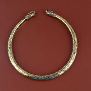 May include: A gold-coloured necklace with detailed floral and scrollwork patterns. The ends of the necklace feature animal head designs. The necklace is displayed against a solid red-brown background.