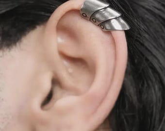 May include: Silver ear cuff with a layered, scale-like design. The cuff wraps around the ear's cartilage, featuring three overlapping segments with small circular details. The jewelry is made of a metallic material.