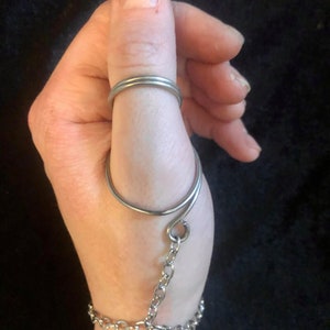 Puede incluir: Un anillo de pulgar de plata con una cadena unida a una pulsera de plata. El anillo está diseñado para envolver el pulgar y la cadena se conecta a una pulsera que se ajusta a la muñeca.