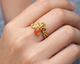 May include: A gold-colored ring featuring a teardrop-shaped, orange gemstone. The ring has an intricate, woven design around the gemstone and band. The ring is a piece of jewelry.