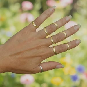 Puede incluir: Una mano que muestra una colección de anillos dorados. Los anillos varían en diseño, algunos rodean todo el dedo y otros forman una banda parcial. Un anillo plateado está en el dedo meñique. El fondo es una vista borrosa de un jardín de flores.