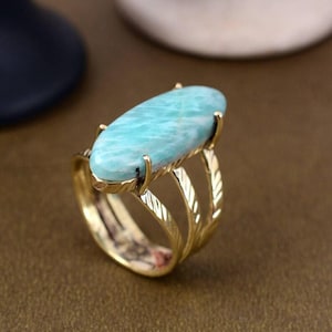Puede incluir: Anillo dorado con una gran gema ovalada de color azul claro. El anillo tiene un diseño multibanda con patrones en forma de hojas en los laterales. La gema está sujeta por cuatro garras.