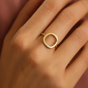 May include: A gold-colored ring with a circular design. The ring features a smooth, polished finish and is worn on a finger. The ring's design is a simple, open circle.