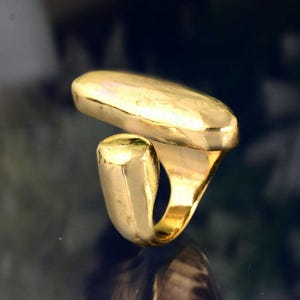 Puede incluir: Un anillo de tono dorado con un diseño abstracto único. El anillo presenta dos piezas conectadas, una ovalada redondeada y la otra de forma curva y alargada.