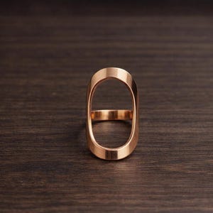 May include: A rose gold ring with an elongated oval design. The ring has a smooth, polished finish and a minimalist aesthetic. The ring is set against a dark wood grain background.