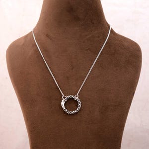 May include: A silver-colored necklace featuring a circular pendant with a textured snake-like design. The pendant is connected to a delicate chain, displayed on a brown mannequin. The necklace is a piece of jewelry.