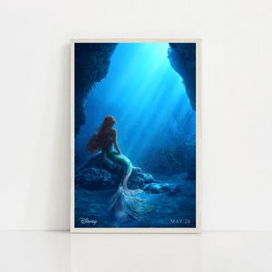 Puede incluir: Una impresi&oacute;n enmarcada que presenta una sirena con largo cabello rojo, una cola verde y un top de concha, sentada en una roca bajo el agua. El fondo es azul vibrante con rayos de luz. El logotipo de Disney y la fecha "MAY 26" son visibles.