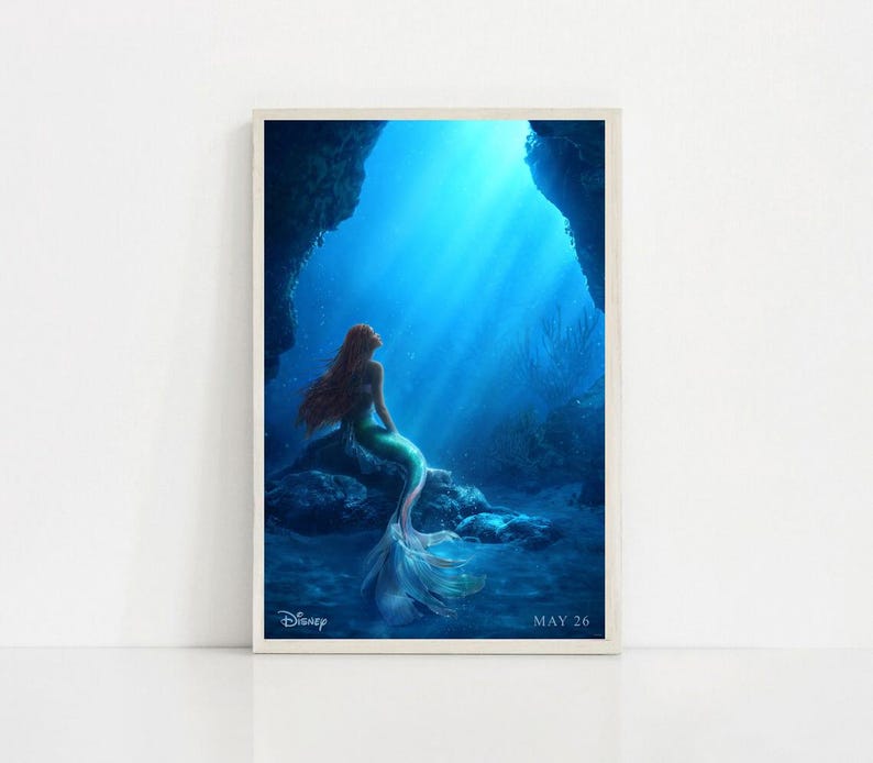 Puede incluir: Una impresi&oacute;n enmarcada que presenta una sirena con largo cabello rojo, una cola verde y un top de concha, sentada en una roca bajo el agua. El fondo es azul vibrante con rayos de luz. El logotipo de Disney y la fecha "MAY 26" son visibles.