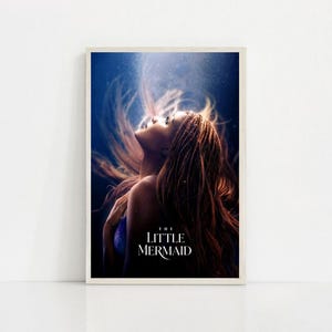 Puede incluir: Un p&oacute;ster enmarcado que presenta a una mujer con largo cabello casta&ntilde;o rojizo que fluye, mirando hacia arriba. La imagen tiene un tema submarino azul y blanco, con el texto "THE LITTLE MERMAID" en blanco en la parte inferior. El p&oacute;ster est&aacute; sobre un fondo blanco.