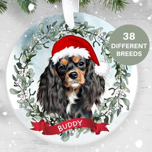 May include: Round Christmas ornament featuring a Cavalier King Charles Spaniel wearing a Santa hat. The dog is surrounded by a wreath of greenery and berries. The name "Buddy" is on a red banner. Text on the ornament reads "38 Different Breeds".