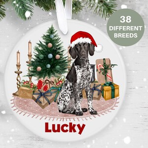 May include: A round, white Christmas bauble featuring a black and white German Shorthaired Pointer dog wearing a red Santa hat. The dog is sitting in front of a small Christmas tree with presents and candles. The bauble has the name "Lucky" written on it.