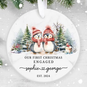 May include: A white ceramic bauble with a watercolour illustration of two penguins wearing red hats and scarves in a snowy forest. The text reads "Our First Christmas Engaged Sophie ♥ George EST. 2024".