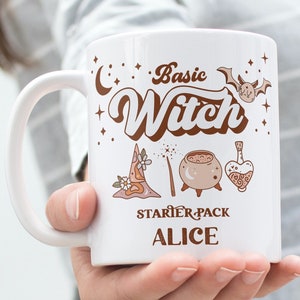 Może przedstawiać: Biały kubek ceramiczny z brązowym wzorem, na którym widnieje napis "Basic Witch Starter Pack Alice". Wzór zawiera półksiężyc, kapelusz czarownicy, kociołek, różdżkę i butelkę z eliksirem.