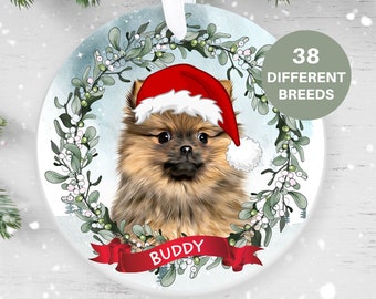 May include: A round, white Christmas ornament with a watercolor background featuring a small, brown and tan Pomeranian dog wearing a red Santa hat. The dog is surrounded by a green wreath with white berries. The ornament has a red ribbon with the name "BUDDY" written on it.