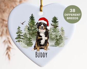 Könnte beinhalten: Weißer, herzförmiger Weihnachtsbaumschmuck aus Keramik mit einer Aquarell-Illustration eines Berner Sennenhundes, der einen Weihnachtsmann-Hut in einem verschneiten Wald trägt. Der Anhänger hat den Text "BUDDY" unter dem Hund.