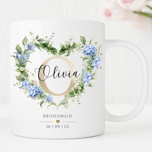Op de afbeelding: Witte keramische mok met een gouden hartvormig bloemendesign met blauwe bloemen en groene bladeren. De naam "Olivia" staat in zwarte schrift in het hart. De tekst "BRIDESMAID" en "16 | 09 | 25" staat onder het hart.