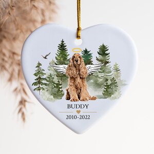Peut inclure: Ornement en céramique blanche en forme de cœur avec une illustration aquarelle d'un cocker spaniel avec des ailes d'ange dans un décor de forêt. L'ornement porte l'inscription "BUDDY" et "2010-2022".