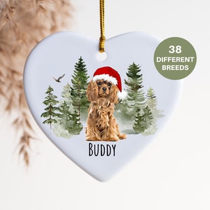 Op de afbeelding: Hartvormige kerstversiering met een Cavalier King Charles Spaniel met een kerstmuts, tegen een bosachtergrond. De naam van de hond, "Buddy", staat eronder gedrukt. Een cirkelvormig label leest "38 VERSCHILLENDE RASSEN."