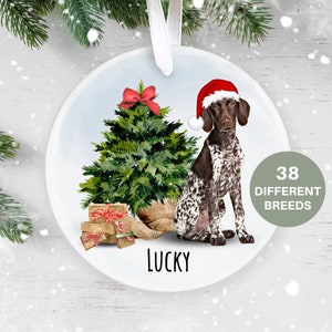 May include: A round, white Christmas bauble featuring a German Shorthaired Pointer dog wearing a red Santa hat, sitting next to a green Christmas tree with presents underneath. The bauble has the word "LUCKY" printed on the bottom.