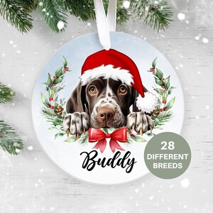 May include: A round Christmas ornament featuring a brown and white German Shorthaired Pointer dog wearing a red Santa hat. The dog is peeking out from behind a wreath of green leaves and red berries. The ornament is personalized with the name "Buddy" and the text "28 Different Breeds".