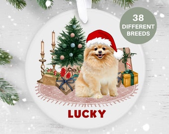 Könnte beinhalten: Ein weißer Keramik-Weihnachtsbaumschmuck mit einem Zwergspitz, der einen Weihnachtsmann-Hut trägt und vor einem Weihnachtsbaum mit Geschenken und Kerzen sitzt. Der Schmuck ist mit dem Namen "LUCKY" personalisiert.