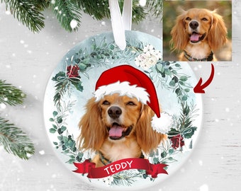 May include: A round, white ceramic ornament with a watercolor-style design of a dog wearing a Santa hat. The dog is surrounded by greenery and white flowers. The ornament has the word "TEDDY" written on a red ribbon.