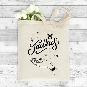 May include: A white canvas tote bag with black lettering that says "Taurus" with a Taurus zodiac symbol above the word. The bag has a black outline of a hand with stars around it.