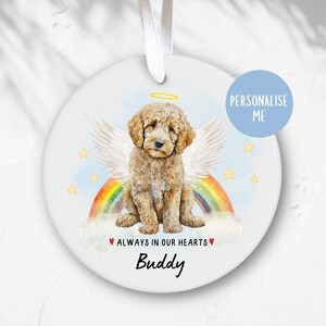 Op de afbeelding: Een witte keramische ornament met een aquarel-illustratie van een bruine hond met engelenvleugels die op een regenboog boven wolken zit. De tekst "ALWAYS IN OUR HEARTS" is in rood geschreven met een hart aan elke kant. De naam "Buddy" staat onder de tekst.