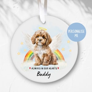 Op de afbeelding: Witte keramische hanger met een aquarel-illustratie van een bruine en witte hond met engelenvleugels en een halo. De hond zit op een wolk met een regenboog erachter. De tekst "ALWAYS IN OUR HEARTS" en "Buddy" staat onder de hond.