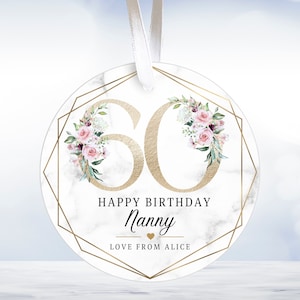May include: Round white marble ornament with a gold geometric frame and pink floral design. The number 60 is in gold inside the frame. The text "Happy Birthday Nanny Love from Alice" is below the number.