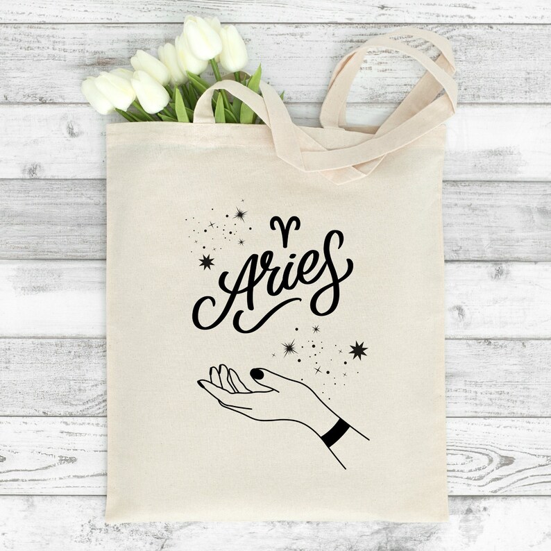 May include: A white canvas tote bag with black text and a graphic. The text reads "Aries" with a stylized ram symbol above it. A hand with a bracelet is reaching up towards a cluster of stars.