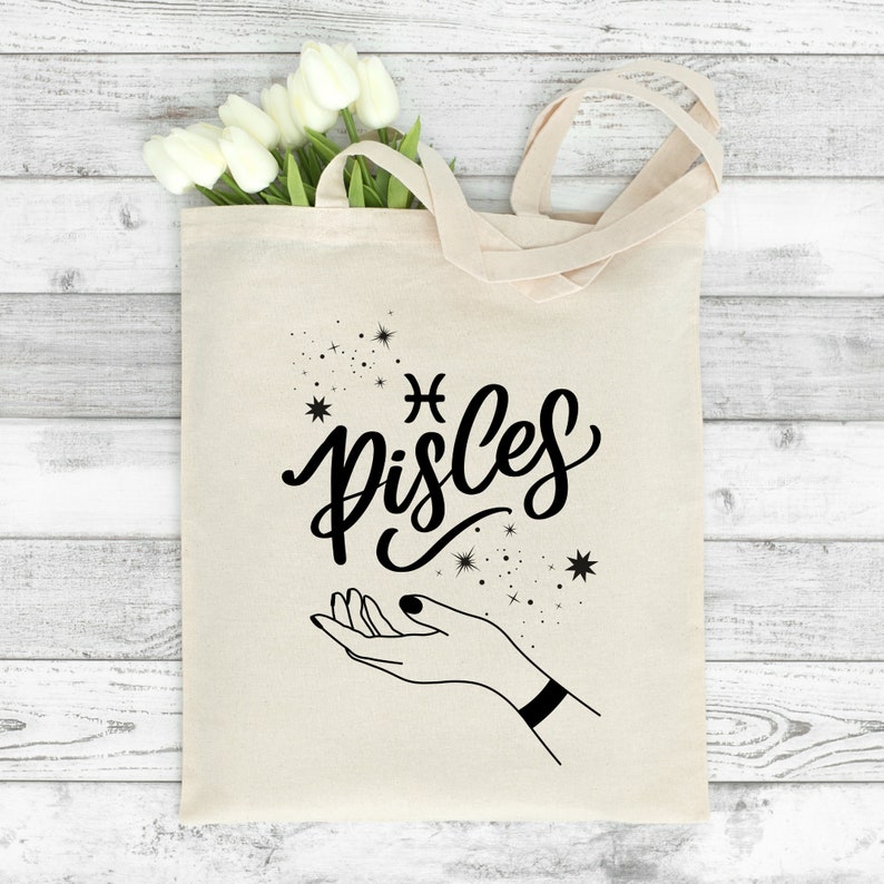 May include: A white canvas tote bag with black text that says "Pisces" and a stylized Pisces astrological symbol. The bag also features a black line drawing of a hand with stars around it.