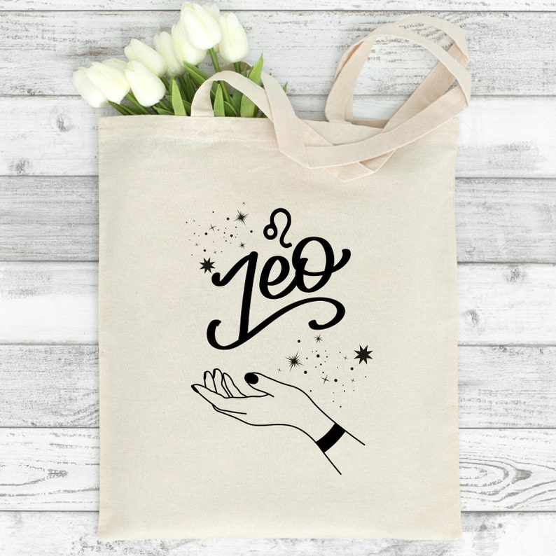 May include: A white canvas tote bag with black text and a graphic design. The text says "Leo" with the astrological symbol for Leo above it. The graphic is a hand with stars around it.