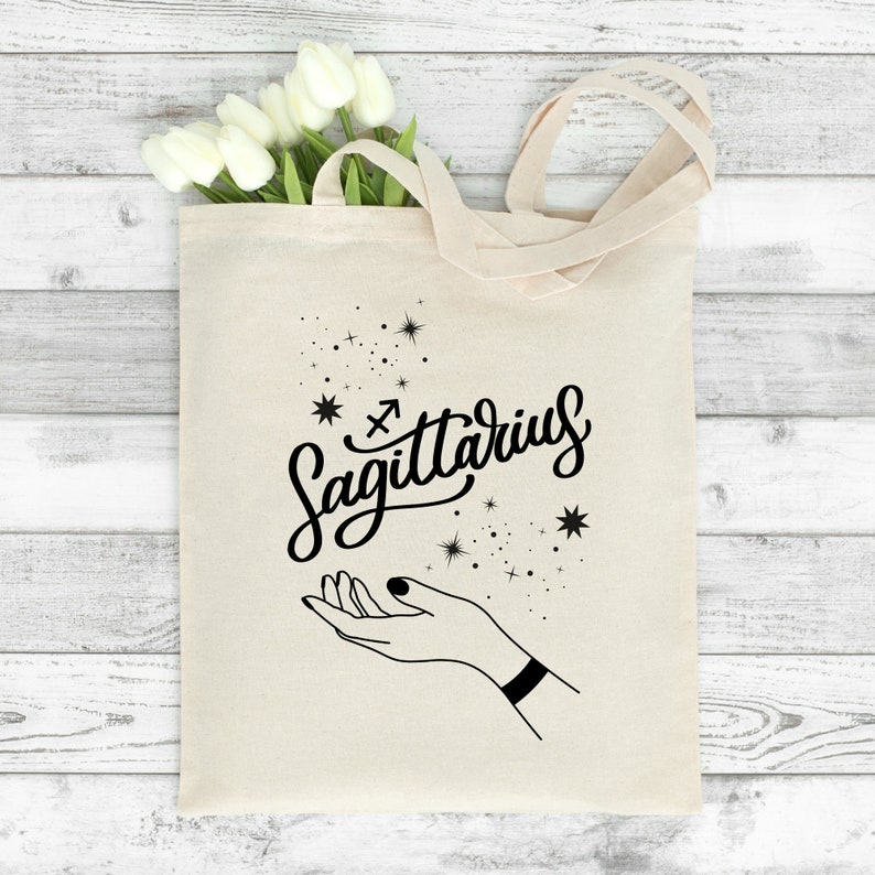 May include: A white canvas tote bag with black text and a graphic. The text says "Sagittarius" with a Sagittarius zodiac symbol. The graphic is a hand with stars around it.