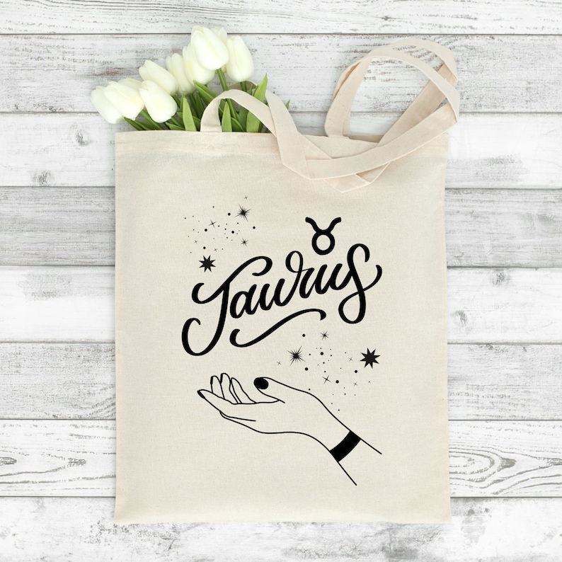 May include: A white canvas tote bag with black lettering that says "Taurus" with a Taurus zodiac symbol above the word. The bag has a black outline of a hand with stars around it.