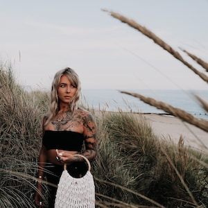 May include: A woman with tattoos on her chest and arms stands in a field of tall grass. She is wearing a black strapless top and holding a white crocheted bag with a wooden handle.