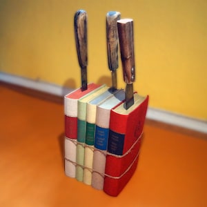Könnte beinhalten: Ein Stapel von fünf Büchern, gebunden in verschiedenen Farben, darunter Rot, Grün, Blau und Creme, mit drei Messern mit Holzgriffen, die in die Oberseite des Stapels gesteckt sind.
