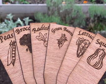 May include: A collection of wooden garden markers, each engraved with a different vegetable name and illustration. The markers include peas, broccoli, radish, green beans, lettuce, and squash. The background shows a garden bed.