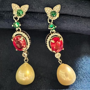 Könnte beinhalten: Ein Paar goldene Ohrhänger mit einem schmetterlingsförmigen Oberteil, besetzt mit kleinen klaren Steinen und einem grünen Edelstein. Darunter befinden sich ein ovaler roter Edelstein mit klaren Steinen und eine cremefarbene Tropfenperle.