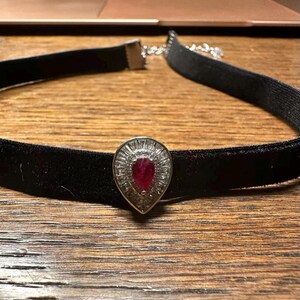 Könnte beinhalten: Ein schwarzes Samt-Halsband mit einem zentralen tropfenförmigen Anhänger. Der Anhänger enthält einen tiefroten, birnenförmigen Edelstein, der von einem Kranz aus kleinen, klaren Baguette-geschliffenen Steinen in Silber gefasst ist.