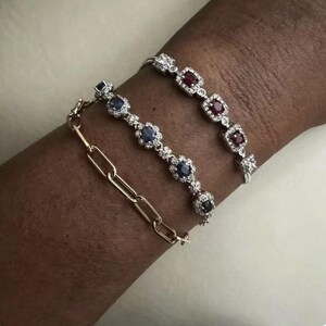 Peut inclure: Trois bracelets sur un poignet. L'un est un bracelet à maillons en or. Les deux autres sont des bracelets de pierres précieuses, l'un avec des pierres bleues et l'autre avec des pierres rouges, le tout serti d'argent avec de petites pierres d'accent.