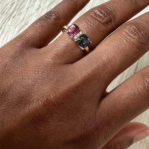 Könnte beinhalten: Ein goldener Ring mit drei quadratischen Edelsteinen: ein rosa, ein blauer und eine Ansammlung kleiner weißer Steine. Der Ring befindet sich an einer Hand, mit hellem Hintergrund.